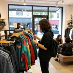 Urban shopping scene with a shopper examining a colorful jacket and smartphone near accessories, inspired by https://forumup.org/shop/search.php?ca_id=&addr1=&addr2=&addr3=&schKeyword=.